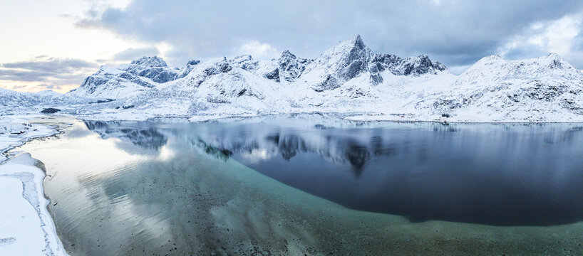 Aerial view of jagged, snow-covered peaks mirrored in the icy waters, the shoreline dusted with white, creating a serene winter landscape, Flakstadpollen, Lofoten, Norway.