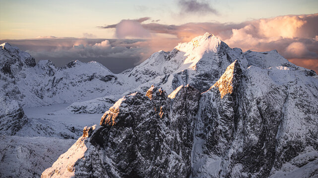 Aerial view of jagged peaks kissed by the golden light of dawn, snow-dusted slopes contrasting with the dark, rocky terrain, Lofoten, Norway.
