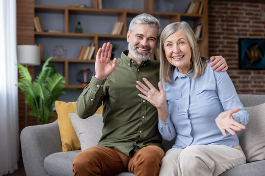 Mature smiling couple at home waving and talking on a live webcam video call, connecting with family and friends for joyful remote conversation and digital togetherness - Powered by Adobe