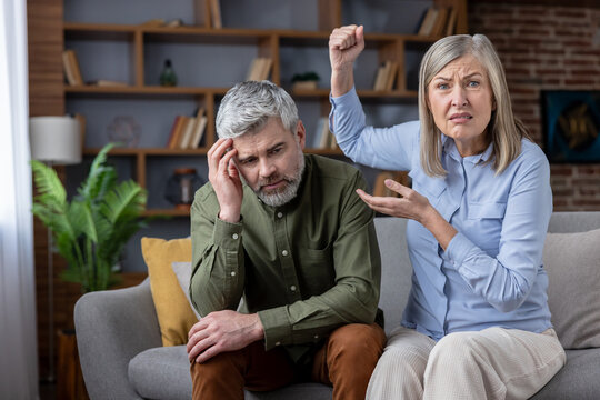Angry senior woman shouting and gesturing aggressively at her sad husband, who is sitting on the sofa feeling stressed and frustrated during a domestic dispute
