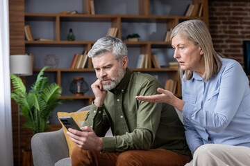 Mature couple having a disagreement while the man is distracted by his smartphone, highlighting issues of neglect and communication problems in their relationship at home