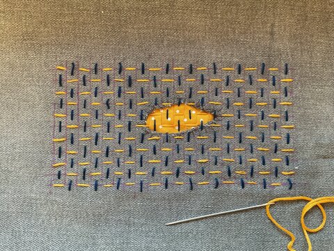 This delicate Sashiko repair transforms a torn hole into an eye-shaped focal point, elegantly stitched with precise running stitches in deep navy and warm golden-yellow thread on a textured fabric