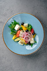 Classic Greek salad with feta cheese cubes, black olives, cherry tomatoes, cucumber, red onion rings, bell pepper and fresh greens in blue bowl. Vibrant Mediterranean appetizer, healthy gourmet dish