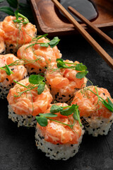 Creamy salmon sushi rolls topped with rich salmon tartare and fresh microgreens, served with soy sauce and chopsticks on dark slate. Luxury Japanese maki, gourmet seafood roll, creamy salmon sushi.