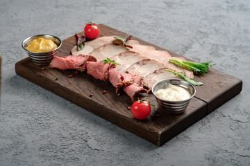 Thinly sliced rare roast beef and tender pork served with creamy horseradish, Dijon mustard, tomatoes and arugula on wooden cutting board. Premium cold cuts assortment, sophisticated meat dish
