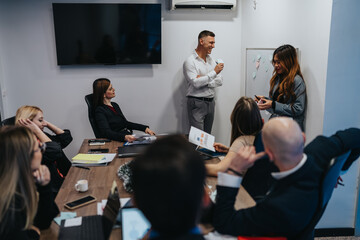 A group of professionals sit around a wooden conference table as a presenter speaks and colleagues share notes. The scene captures a collaborative, modern office moment with teams discussing ideas.