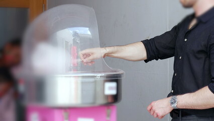 Man spinning cotton candy inside clear dome machine at event or party, vendor preparing treat in...