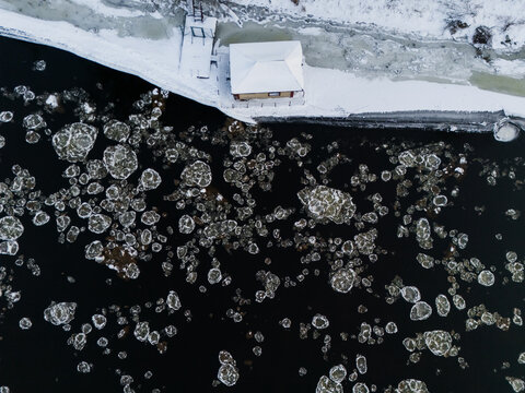 Aerial view of ice floes scattered across the dark river contrasting with the snow-covered banks near a small building, Vilnius, Vilnius County, Lithuania.