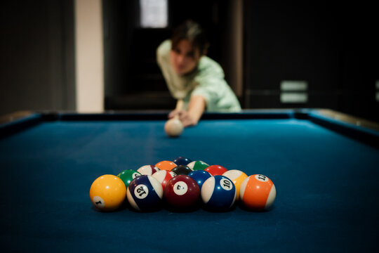 Selective focus image of a billiards table with a racked set of colorful pool balls in the foreground and a player aiming the cue ball in the background. 