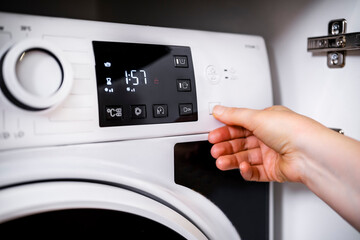 Housewife starts washing machine cycle by pressing start button. Modern white laundry appliance in...
