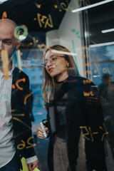Two professionals in a modern office discuss ideas while writing on a glass wall filled with colorful notes. A collaborative moment in a contemporary workspace shows teamwork and planning.