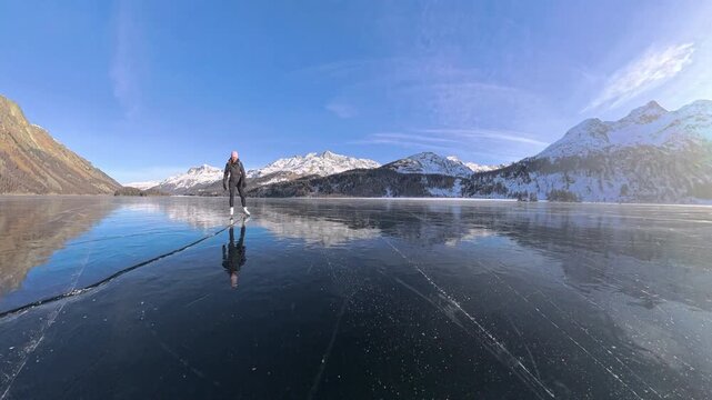 Woman ice skating across mirror-like black ice on frozen Sils Lake with reflections of the Swiss Alps