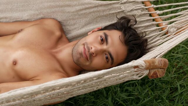 Opening eyes from sunlight, shirtless man shifting gaze and smiling in garden hammock with spreader