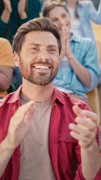 Caucasian handsome young male laughing while celebrating football moment in stadium crowd. Cheerful man in red shirt smiling wide surrounded by clapping fans. Excited reaction during game.