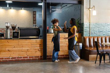 Two female baristas wearing black aprons standing and gesturing in cafe at counter by espresso