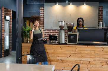 Female baristas in aprons using tablet and phone, operating espresso machine at coffee shop counter