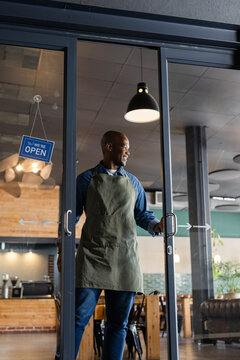 African American man standing holding glass door handle at cafe entrance wearing olive green apron