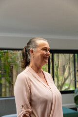 Mature woman standing by black windows in living room, wearing pale pink blouse, teal sofa visible