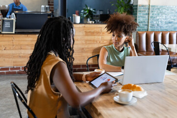 African American colleagues wearing sleeveless tops discussing data with laptop and coffee at cafe