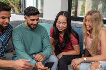 Diverse friends sitting on gray couch in living room, sharing smartphone and pointing at screen