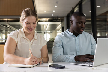 Diverse coworkers writing and typing at white table in open-plan office with laptop notebook phone