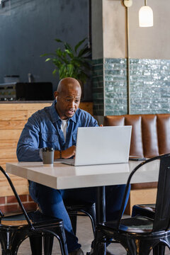 African American man sitting at cafe, working on silver laptop with earbuds, takeaway cup, phone