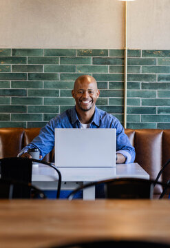 African American man 30s denim-shirt sitting at white table in cafe holding laptop and togo cup