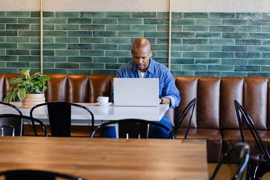 African American adult man sitting in quiet cafe booth, working on silver laptop with coffee cup