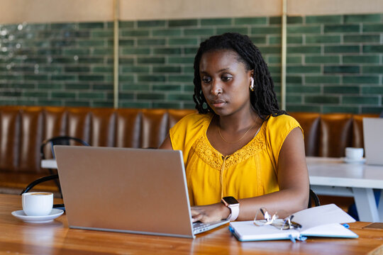 African woman typing on silver laptop in cafe, wearing yellow blouse, notebook and cup nearby