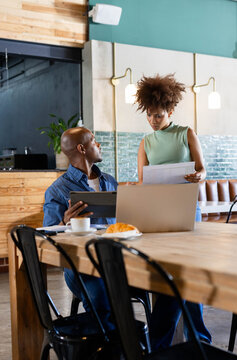 African American coworkers discussing while holding printed documents by laptop and tablet at cafe