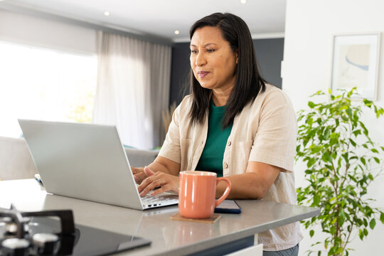 Mature Asian woman typing on laptop at kitchen counter, wearing green top, mug hiding smartphone