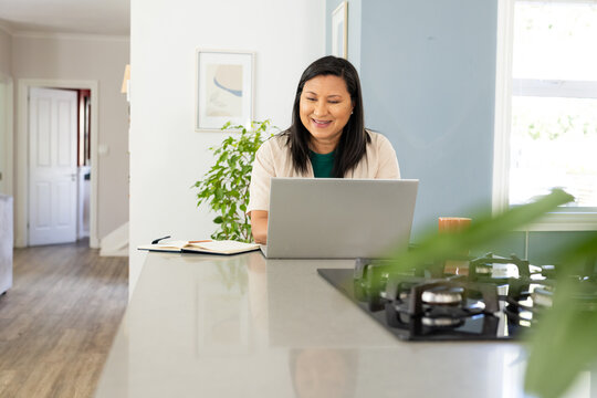 Mature Asian woman working on laptop at kitchen island with notebook pen mug and cooktop