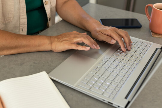Hands are typing on silver laptop with backlit keys on gray countertop, coral mug nearby