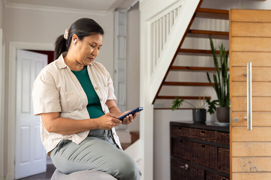 Asian woman sitting on ottoman at home entry holding smartphone, wearing scrunchie and earbud