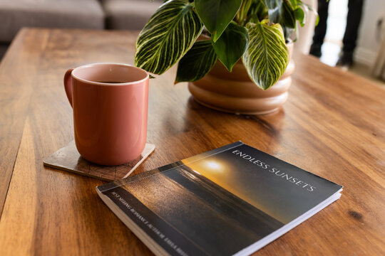 Pink ceramic mug is resting on stone coaster on wooden table with ENDLESS SUNSETS book, plant
