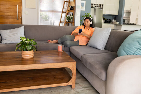 Senior woman lying on sofa in living room wearing orange shirt headphones holding phone, copy space