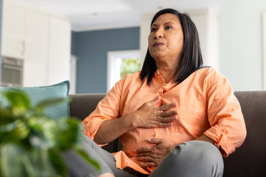 Woman wearing orange button-up shirt and gray pants, clutching chest and abdomen on sofa at home