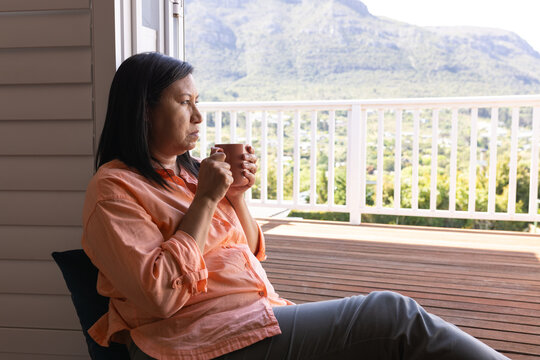 Woman sitting on porch wearing peach shirt gray pants holding ceramic mug, gazing toward valley