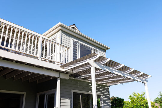 Second-story deck is extending in suburban backyard, with white pergola, glass doors, string lights