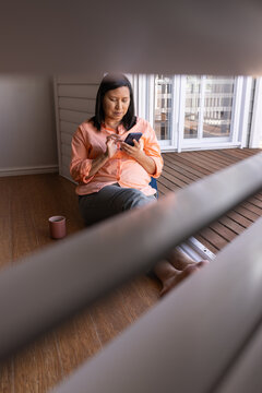 Mature woman sitting on porch wearing peach shirt, gray pants, holding smartphone near ceramic mug