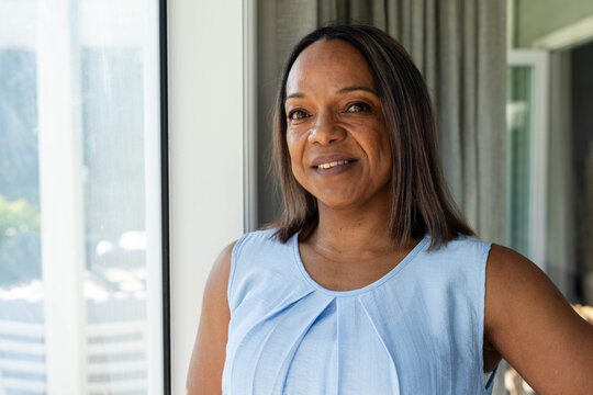 Mature woman standing near large glass window at home with curtains, wearing sleeveless blue blouse