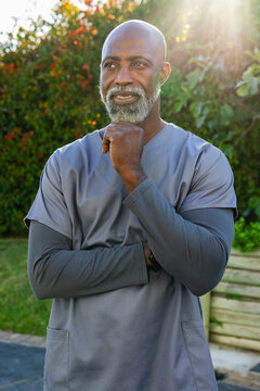 Senior African American man standing in garden wearing gray scrubs and wristwatch, sun flaring