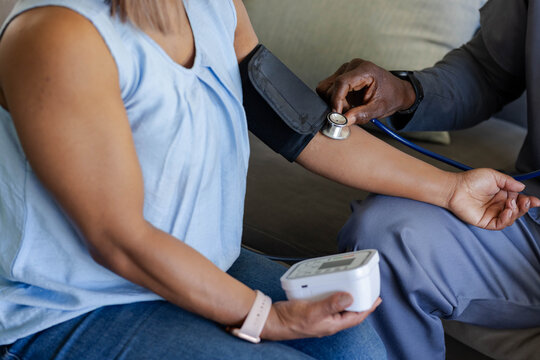 African American man checking BP with stethoscope and cuff while woman holding monitor on sofa