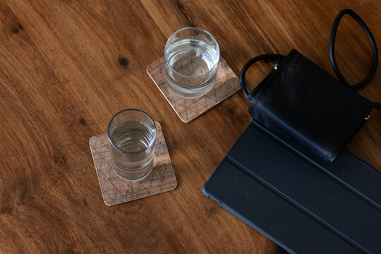 Two clear glasses are sitting on patterned coasters on wooden tabletop, black bag and tablet nearby