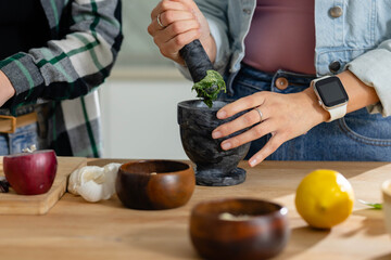 Diverse female friends crushing green herbs in stone mortar on home kitchen counter, wearing denim