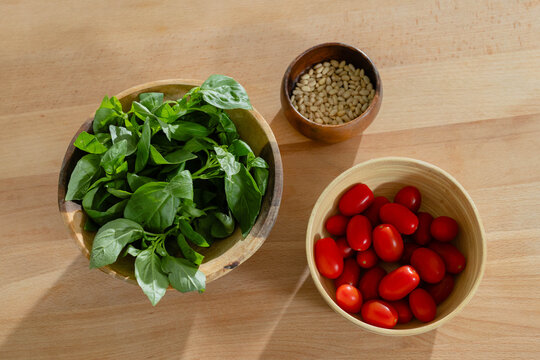 Large wooden bowl of spinach is sitting on light wooden table, with tomatoes and pine nuts
