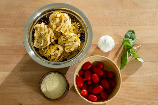 Steel colander is holding three pasta nests on wooden board with tomatoes, cheese, garlic and basil