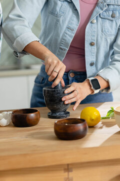 Dark stone mortar and pestle is crushing garlic and herbs on wooden counter near lemon