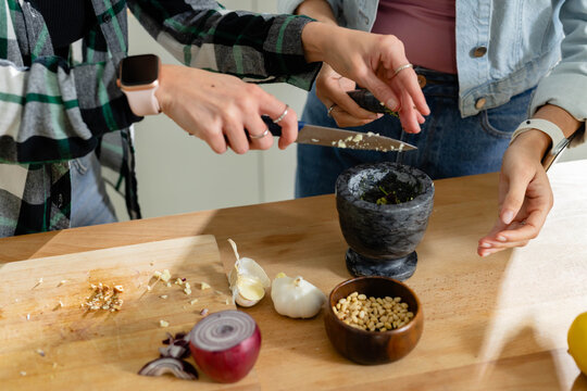 Two adult women preparing garlic at kitchen counter with mortar and chef's knife, wearing denim