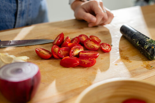 Halved cherry tomatoes resting on wooden cutting board with chef's knife, red onion, muddler